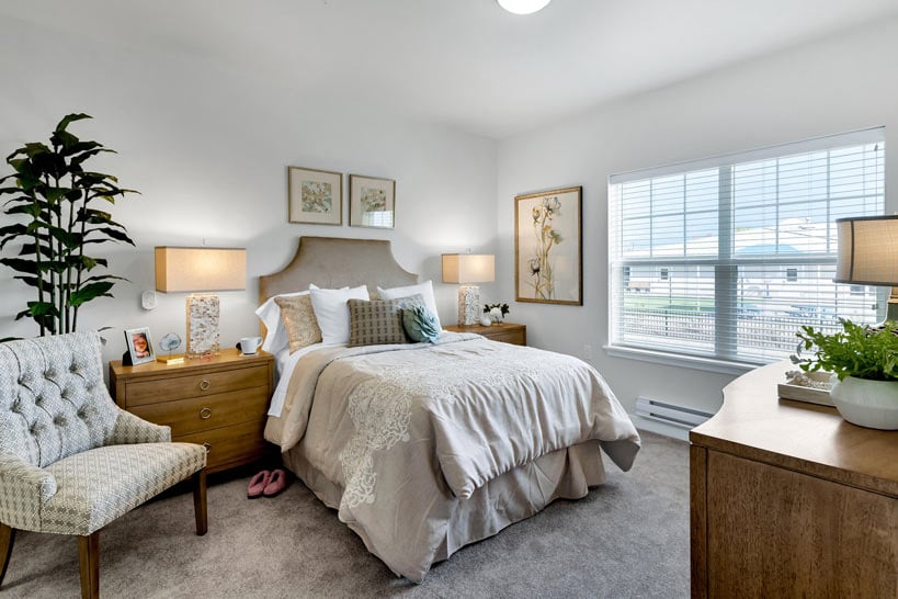A cozy bedroom featuring a queen-sized bed with beige bedding and lace accents. The room includes wooden nightstands with matching lamps, a cushioned chair, plants, and framed wall art. A large window brings in natural light, and a wooden dresser is placed opposite the bed.
