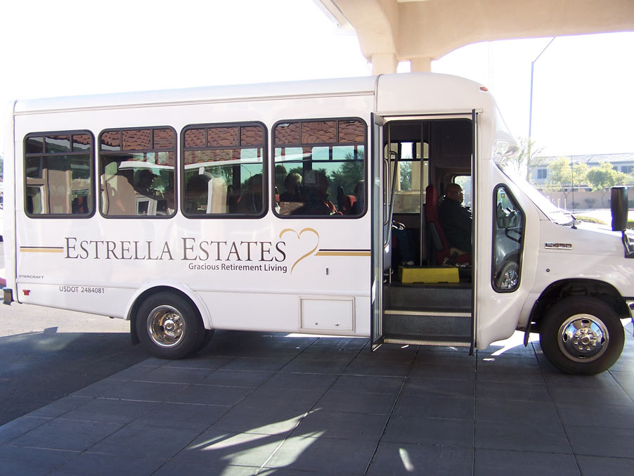 A white shuttle bus with the branding "Estrella Estates - Gracious Retirement Living" is parked under a shaded area. The bus door is open, and a person is seated inside near the entrance. The bus is likely used for transporting residents.