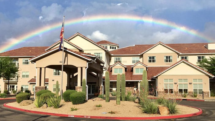 A modern, multi-story building with a beige facade, visible through a landscaped foreground with desert plants like cacti. A rainbow arches across the cloudy sky above the building. An American flag is flying on a flagpole near the entrance.