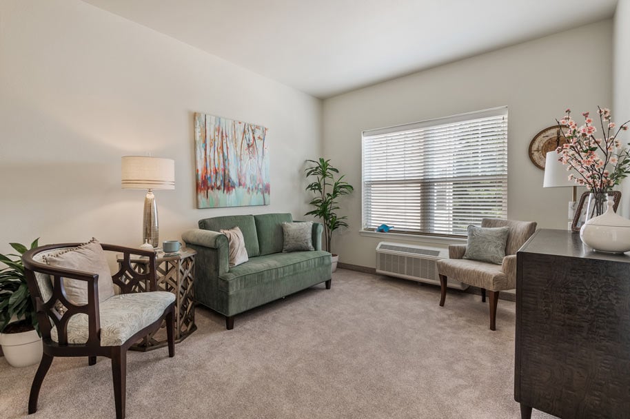 A cozy, well-lit living room with a green sofa, two cushioned chairs, and a wooden desk. The space features a large window with blinds, a floor lamp, various plants, decorative art on the wall, and a vase with pink flowers on the desk. The carpet is a light beige color.