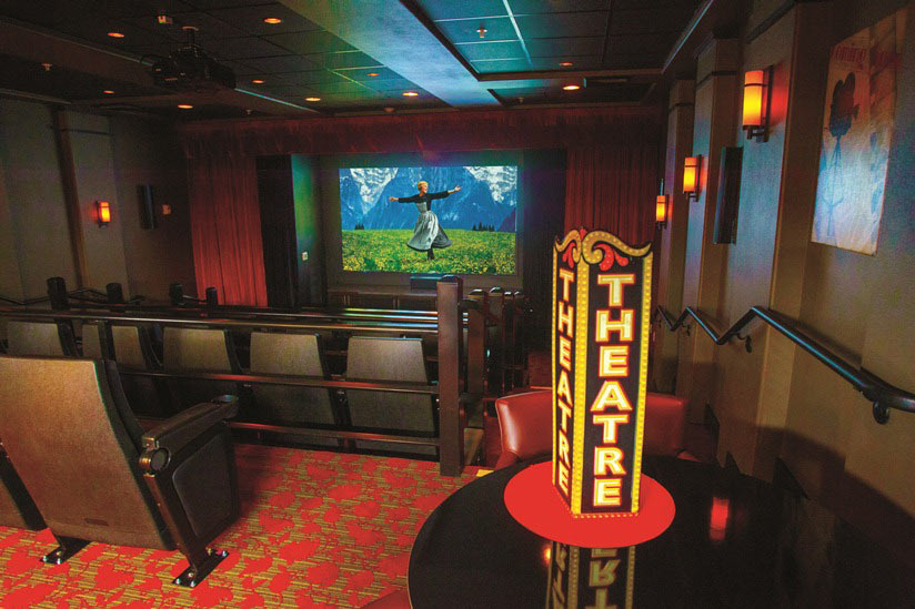 A home theater room with theater seating, red carpet with a patterned design, and red curtains. A brightly lit marquee sign with the word "THEATRE" is placed near the entrance. A movie scene featuring a person in a grassy field with mountains is playing on the screen.