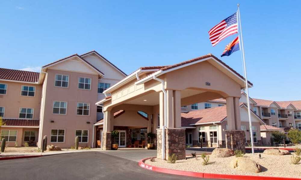 A three-story assisted living facility with a beige exterior and red-tiled roofs. The entrance features a covered driveway, and two flags are flying on flagpoles in the front: the American flag and a state flag. The sky is clear and sunny.